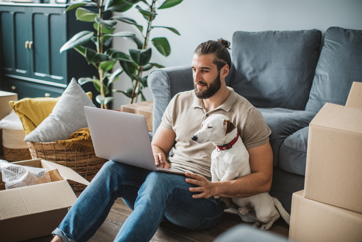 A man with a laptop sits comfortably on the floor against a sofa, accompanied by his dog, in a living room with moving boxes.