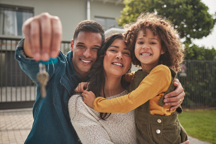 A joyful family with a young child smiles at the camera, with one of the parents holding up a set of keys, suggesting they have just bought a new home.