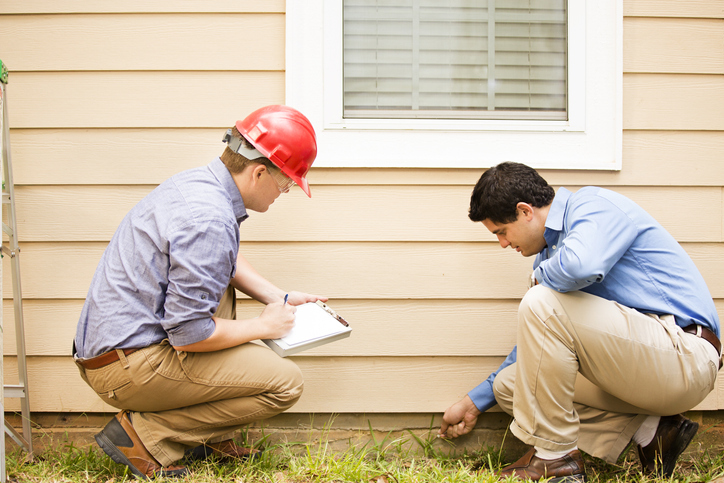 Two men are examining the exterior of a house, with one wearing a red hard hat suggesting that he may be a construction worker, inspector, or engineer. The man with the hard hat is crouched down, taking notes on a clipboard, while the other man is closely inspecting something near the ground.