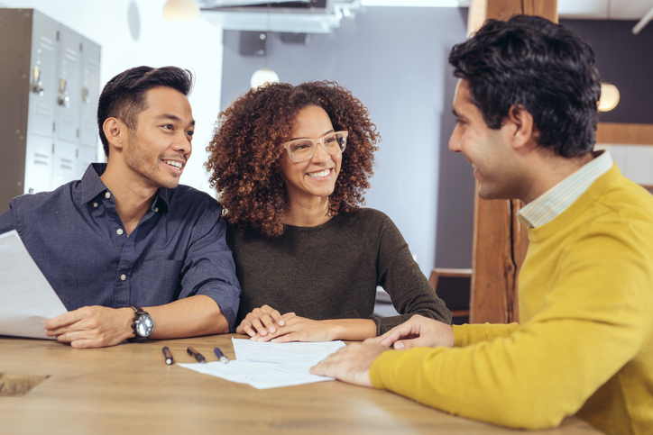 A couple is having a pleasant conversation with a person across the table, with papers in front of them, in what appears to be a meeting or consultation.