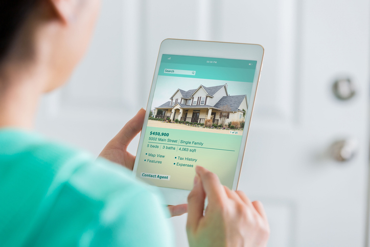 A person holds a tablet displaying a real estate app with a listing for a single-family home, suggesting they are searching for or interested in buying property.