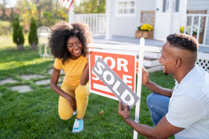 A joyful man and woman are interacting near a "FOR SALE" sign in front of a house. The woman is crouching down and smiling broadly while the man, in a crouched position as well, is attaching a "SOLD" sticker to the sign.