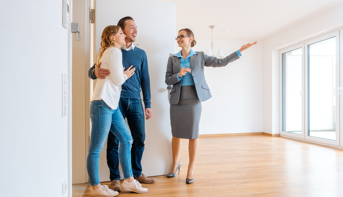 Real estate agent showing a spacious room to a happy couple in a house tour, with the agent gesturing towards large windows with natural light.