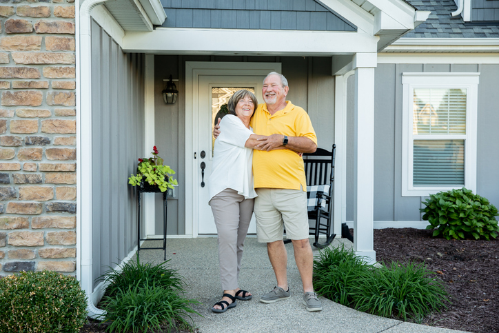 A happy elderly couple embraces in front of their home's entrance, smiling broadly in a welcoming suburban setting.