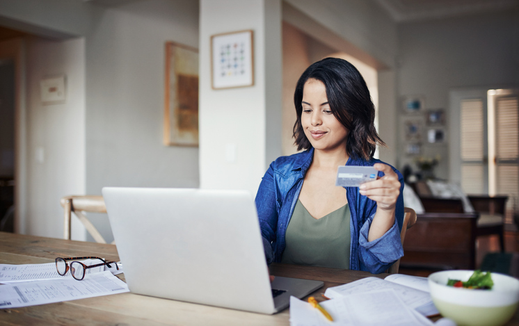 A smiling woman holding a credit card looks at her laptop, surrounded by papers and a pair of glasses on a table in a well-lit home interior.