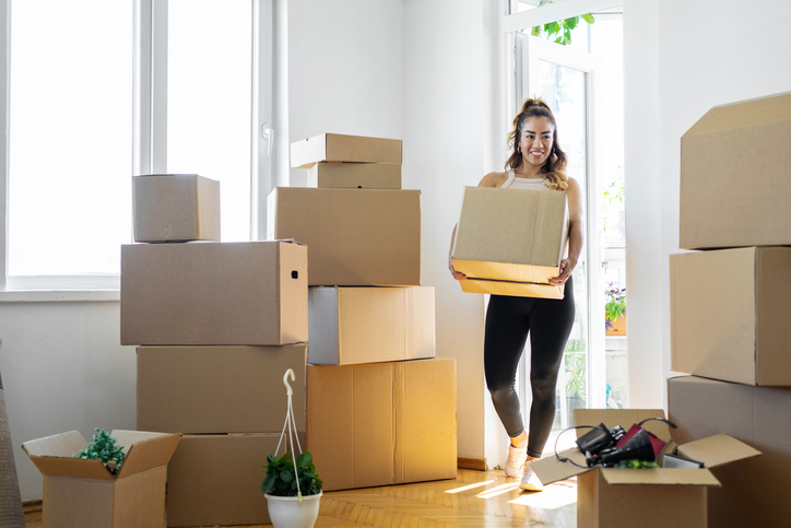 A cheerful woman is carrying a cardboard box in a sunlit room filled with stacked boxes, suggesting a move to a new home. She is wearing a casual outfit suitable for moving day, with a topknot hairstyle, and is smiling, indicating a positive attitude towards the activity.