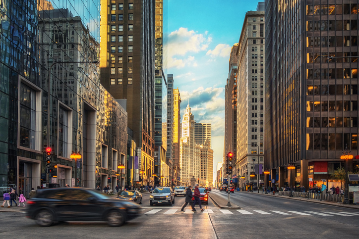 A bustling city street scene with cars in motion and pedestrians crossing, flanked by towering skyscrapers under a sky showing the transition from sunset to evening.