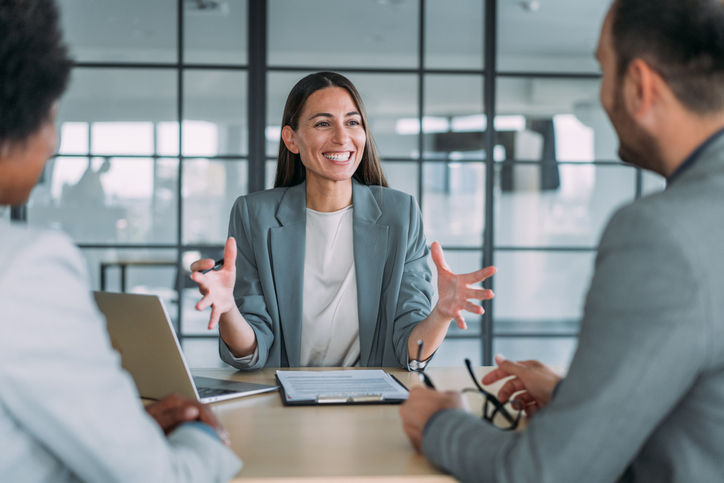 A cheerful professional woman speaks with enthusiasm during a meeting with two other individuals in a bright office with a cityscape visible through the windows behind her.