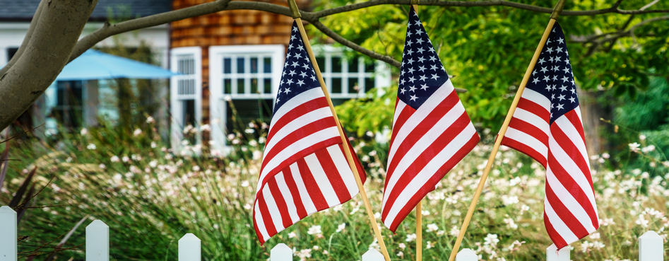 Three American flags hanging on poles, displayed in a residential garden with a white picket fence and a house in the background.